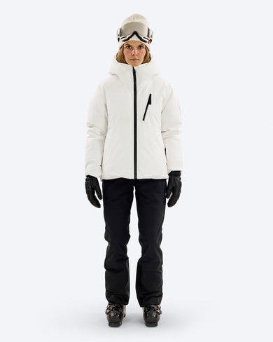 A skier wears The Mountain Studio D-5 W Pertex Reversible Tecno Down Jacket with the matte white side out, featuring a hood, chest pocket, and minimalist design, paired with black ski pants, gloves, ski boots, and goggles for alpine winter use.