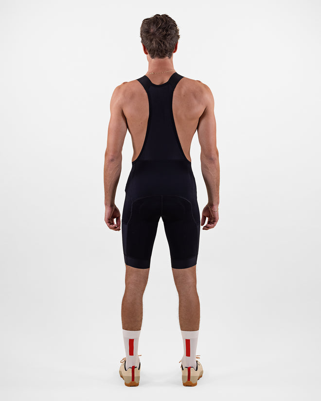 A man is shown from the back wearing The Mountain Studio L-12 men's gravel cycling bib shorts in black, highlighting the ergonomic fit, stretch suspenders, and minimalist design, paired with white socks featuring red stripes and tan cycling shoes.