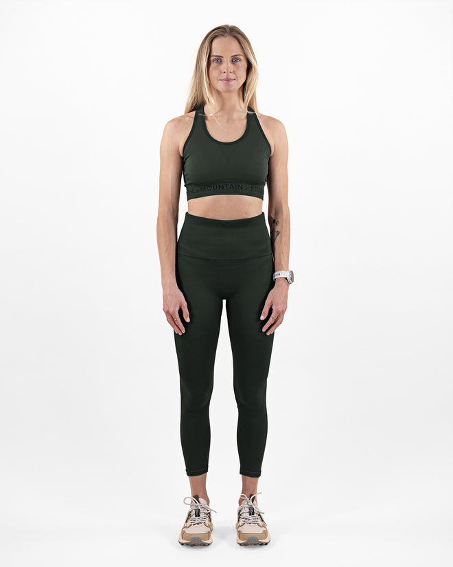Woman wearing The Mountain Studio O-1 W green high-waisted compression leggings with a wide waistband, 7/8 length, and right leg cargo pocket, styled for active outdoor or gym use.