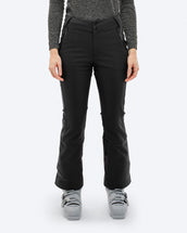 A woman wears The Mountain Studio P-4 W black 2L Stretch Ski Pants, featuring a high waistband, zippered hand pockets, and reinforced lower legs, styled with ski boots and a dark technical top for alpine skiing.