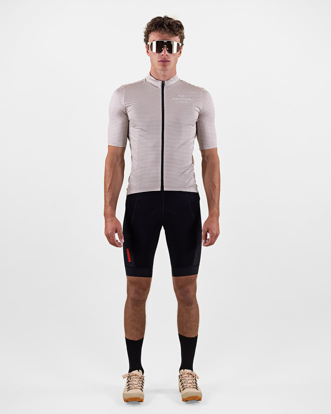 Man wearing The Mountain Studio T-17 Gravel Stripe Jersey in beige with thin horizontal stripes, full-length front zipper, and minimalist logo, paired with black cycling shorts, black socks, and beige cycling shoes, standing against a plain background.