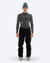 Man wearing The Mountain Studio Y-1 EPE black Gore-Tex ski pants with ergonomic fit, visible ventilation zippers, and reinforced scuff guards, styled with a technical long-sleeve top and ski boots against a plain background.