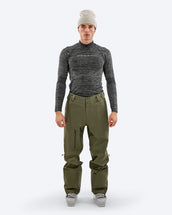 Model wearing The Mountain Studio Y-1 EPE olive green Gore-Tex ski pants with ergonomic fit, zippered leg pocket, and reinforced scuff guards, paired with a technical baselayer and ski boots in a studio setting.