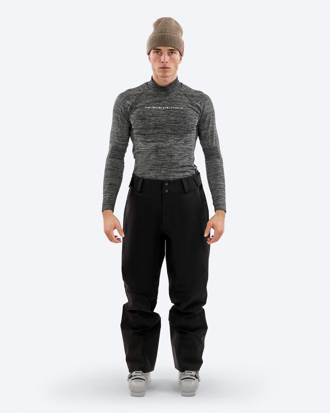 Man wearing The Mountain Studio Y-3 black shell ski pants with articulated knees and reinforced cuffs, paired with a technical long-sleeve top and ski boots, shown in a studio setting.