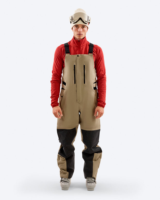 Man wearing The Mountain Studio Y-7 PRO two-tone beige and black Gore-Tex Pro 3-layer ski bib pants with reinforced knees, adjustable suspenders, and zippered chest pockets, styled with a red technical top, ski boots, beanie, and goggles.