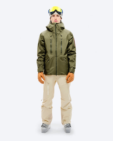 A skier wears the olive green Z-2 EPE shell jacket by The Mountain Studio, featuring a helmet-compatible hood, multiple zippered pockets, and ergonomic fit, paired with beige ski pants, gloves, and ski boots, ready for alpine conditions.