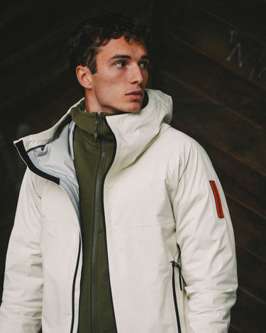Person wearing the Z-4 L shell jacket in white from The Mountain Studio, featuring a minimalist design, adjustable hood, and water-resistant zippers, styled over an olive green midlayer in an outdoor setting.