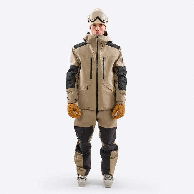 A skier models the Z-7 PRO shell jacket in two-tone beige from The Mountain Studio, featuring reinforced black Vectran panels on the shoulders, elbows, and knees, helmet-compatible hood, and ergonomic alpine fit with ski gloves and boots.