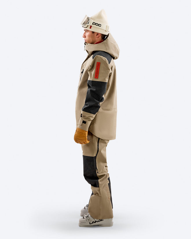 Side profile of a skier wearing the Z-7 PRO shell jacket in two-tone beige from The Mountain Studio, showing reinforced black Vectran panels on the arms and legs, helmet-compatible hood, and ergonomic alpine fit with ski gloves and boots.