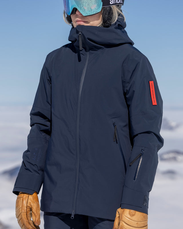 Close-up of a woman wearing The Mountain Studio Z-8 W hybrid shell ski jacket in navy blue, featuring a helmet-compatible hood, waterproof zippers, and ALP-3 membrane, styled for alpine skiing in snowy mountain conditions.