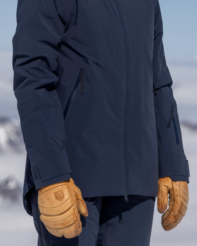 Detail view of The Mountain Studio Z-8 W hybrid shell ski jacket in navy blue, highlighting ALP-3 membrane branding, waterproof zippers, and ergonomic sleeve design, worn with tan ski gloves in a snowy alpine setting.