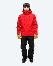 Person dressed for alpine skiing in The Mountain Studio Y-1 PRO GORE-TEX Pro 3L shell pants in black, paired with a red technical jacket, tan gloves, ski boots, and a helmet with goggles, highlighting the pants' ergonomic fit, zippered thigh pocket, and reinforced cuffs.